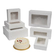 White bakery cake boxes with clear window cutouts, shown in various sizes and configurations on a white background, with a small white frosted cake placed in front.